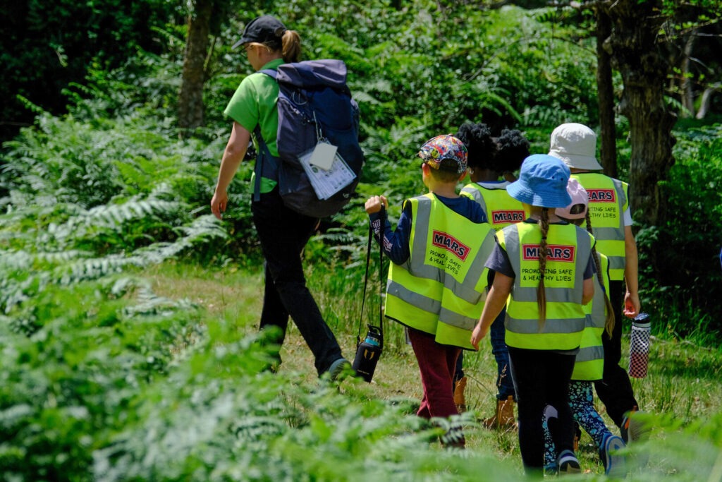 A woman in a green top and rucksack leads a group of schoolchildren on a walk in a woodland setting