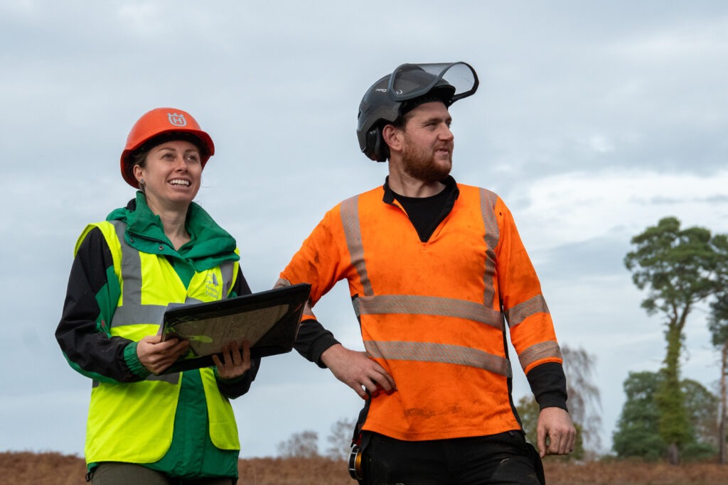 Two workers in high-vis gear and helmets stand outdoors, one holding a clipboard, looking across a wetland restoration site