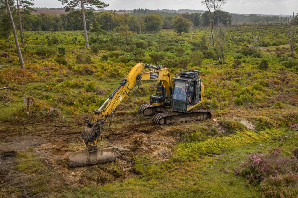Yellow excavator digging a shallow trench in a heathland mire, surrounded by ferns, heather and scattered trees