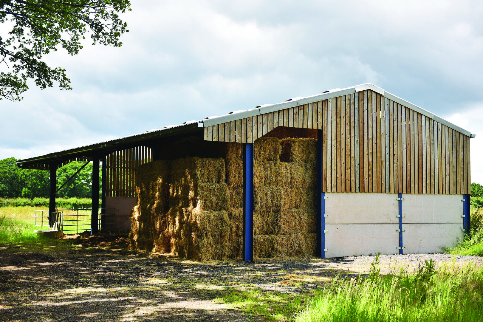 Open-sided wooden barn in the New Forest with stacked hay bales inside, beside a grassy field under a cloudy sky